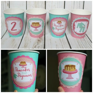 May include: Four paper cups with pink and teal designs. The cups have different designs, including a pancake with syrup, a pair of pajamas, and the number "2". The text "Pancakes and Pajamas" is on two of the cups.
