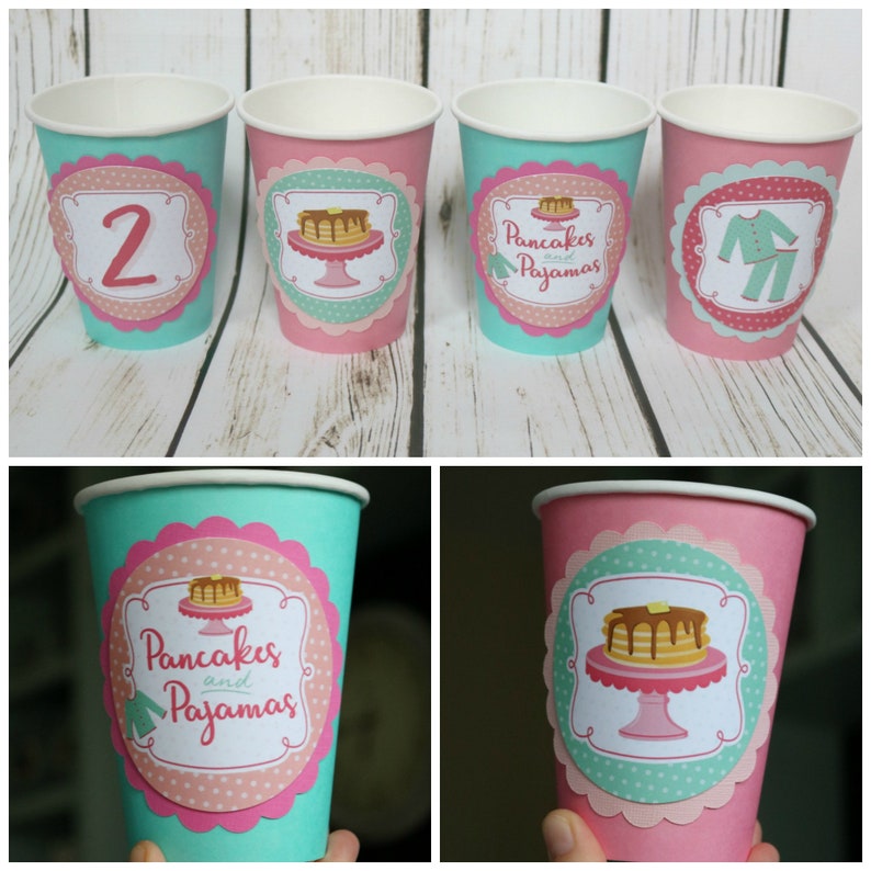May include: Four paper cups with pink and teal designs. The cups have different designs, including a pancake with syrup, a pair of pajamas, and the number "2". The text "Pancakes and Pajamas" is on two of the cups.