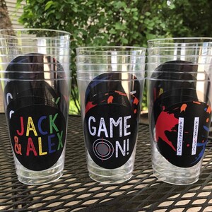 May include: Three clear plastic cups with black and white circular stickers. The stickers have different designs, including the text "Jack & Alex", "Game On!" and a red, blue, and orange paint splatter design.