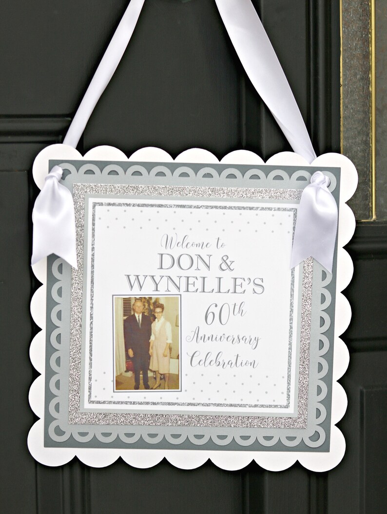May include: A white sign with a scalloped edge and a gray border. The sign has a photo of an older couple, the text "Welcome to Don & Wynnelle's 60th Anniversary Celebration", and a silver glitter background.
