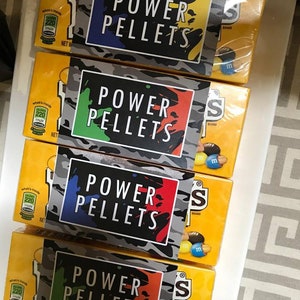 May include: Four boxes of M&M's candy with custom labels that say "Power Pellets" in black text on a camouflage background. Each label has a different color scheme: blue, green, red, and orange.