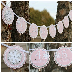 May include: A decorative banner featuring scalloped circular cards in light pink and silver glitter, strung on a white ribbon. Each card displays a number from one to six and the word months, accented with white snowflake cutouts and rhinestones.