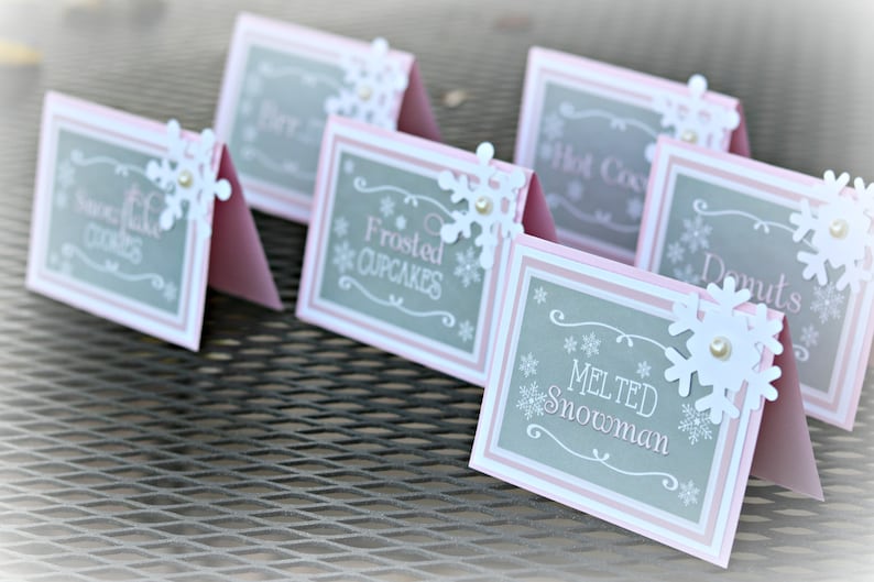 May include: Five pink and gray table cards with snowflake decorations. The cards have the following text: "Sweet Milk Cookies", "Frosted Cupcakes", "Hot Cocoa", "Donuts", and "Melted Snowman".