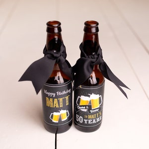 May include: Two brown glass beer bottles with black ribbon bows and personalised labels. The labels are black with gold lettering and a graphic of two beer mugs. The text on the labels reads "Happy Birthday Matt" and "Cheers & Beers to Matt's 50 Years".