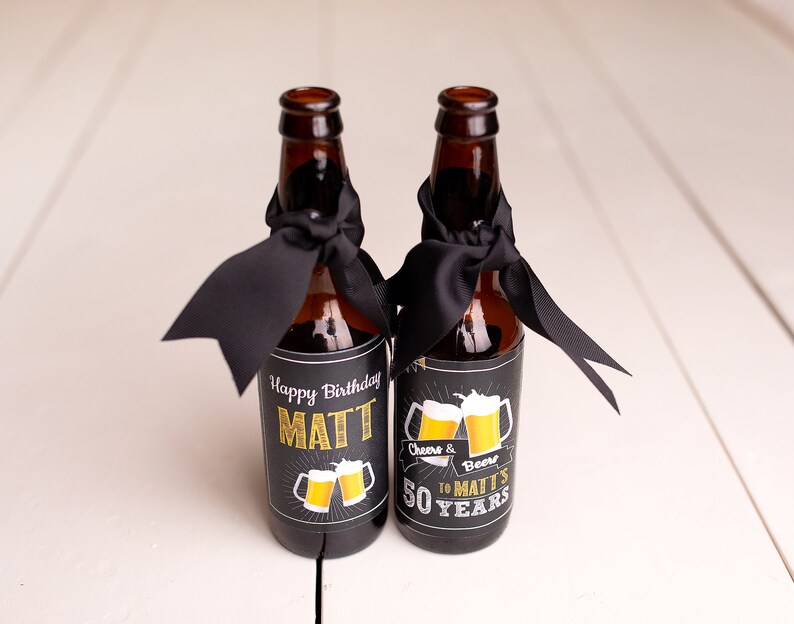 May include: Two brown glass beer bottles with black ribbon bows and personalised labels. The labels are black with gold lettering and a graphic of two beer mugs. The text on the labels reads "Happy Birthday Matt" and "Cheers & Beers to Matt's 50 Years".