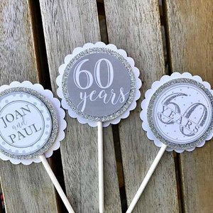 May include: Three silver and white cupcake toppers with scalloped edges. One topper says "60 years" in gray text on a silver background. Another topper says "Joan and Paul" in black text on a white background. The third topper says "60 years" in black text on a white background with silver glitter.