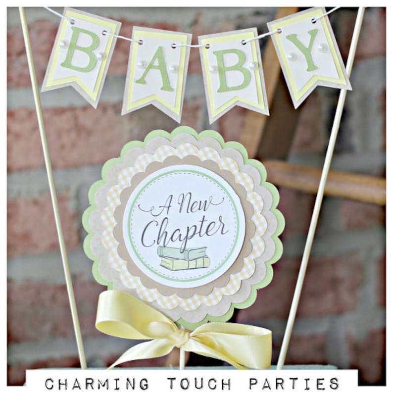 May include: A green and yellow baby shower centerpiece with a banner that reads "BABY" and a circle with the text "A New Chapter" and a stack of books.