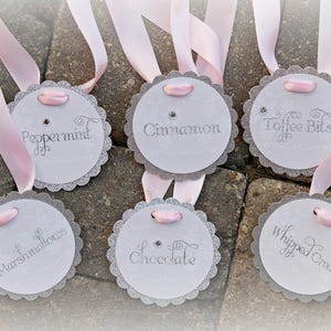 May include: Six circular white tags with scalloped silver glitter edges, each featuring a light pink ribbon loop and a small rhinestone. The tags are printed with the words Peppermint, Cinnamon, Toffee Bits, Marshmallows, Chocolate, and Whipped Cream.