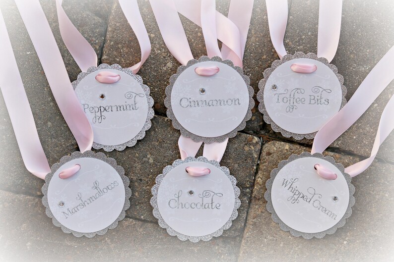 May include: Six circular white tags with scalloped silver glitter edges, each featuring a light pink ribbon loop and a small rhinestone. The tags are printed with the words Peppermint, Cinnamon, Toffee Bits, Marshmallows, Chocolate, and Whipped Cream.