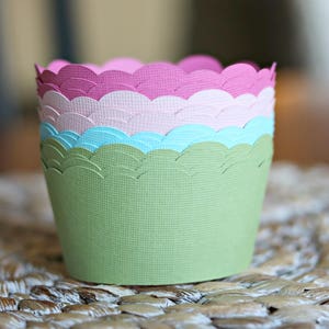 May include: A stack of colorful paper cupcake liners. The liners are in shades of green, blue, pink, and purple, with a scalloped edge design. The liners are stacked on top of each other, creating a visually appealing display.