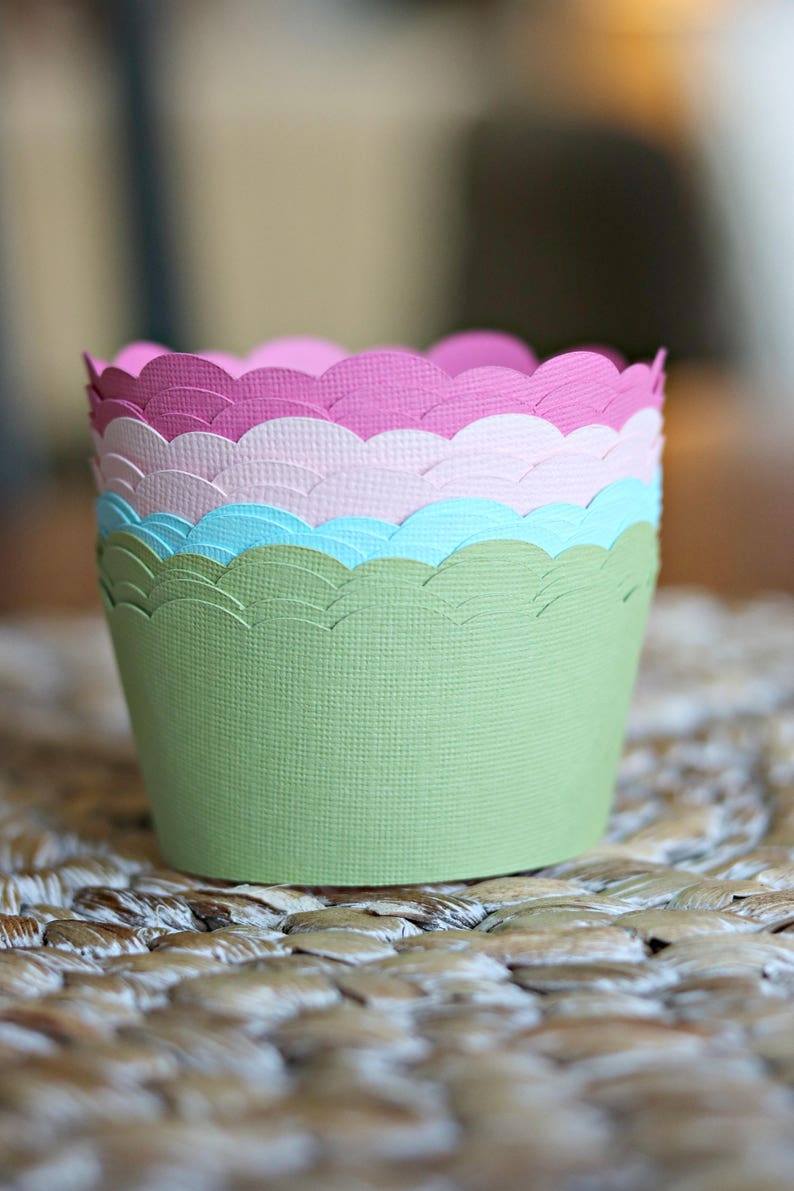 May include: A stack of colorful paper cupcake liners. The liners are in shades of green, blue, pink, and purple, with a scalloped edge design. The liners are stacked on top of each other, creating a visually appealing display.
