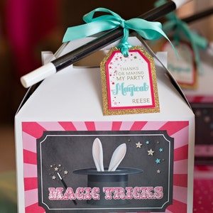 May include: A white favor box with a pink and white striped design. The box has a black and white illustration of a magic hat with bunny ears sticking out. The text "MAGIC TRICKS" is printed in red on the box. A gold tag with the text "THANKS FOR MAKING MY PARTY MAGICAL REESE" is attached to the box with a turquoise ribbon and a black magic wand.