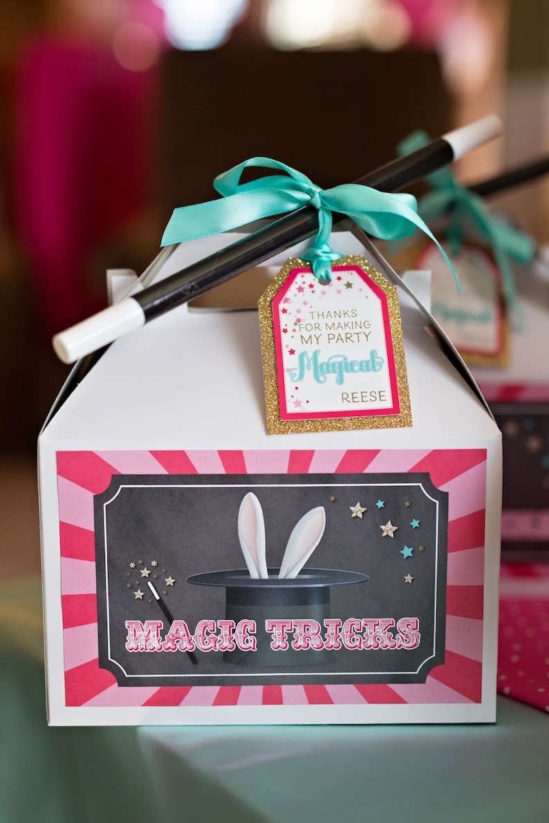 May include: A white favor box with a pink and white striped design. The box has a black and white illustration of a magic hat with bunny ears sticking out. The text "MAGIC TRICKS" is printed in red on the box. A gold tag with the text "THANKS FOR MAKING MY PARTY MAGICAL REESE" is attached to the box with a turquoise ribbon and a black magic wand.