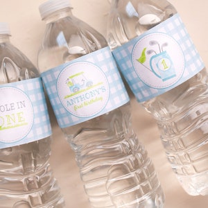 Puede incluir: Tres botellas de agua de pl&aacute;stico transparente con etiquetas de cuadros azules y blancos. Las etiquetas tienen un carrito de golf y palos de golf con el texto "Anthony's first birthday" en una botella y "Hole in One" en otra botella.