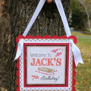 May include: A square sign with a white ribbon hanger featuring a red scalloped border and a white decorative edge. The center displays the text Welcome to JACK'S 7th Birthday! with illustrations of red mittens and a wooden sled on a light background.