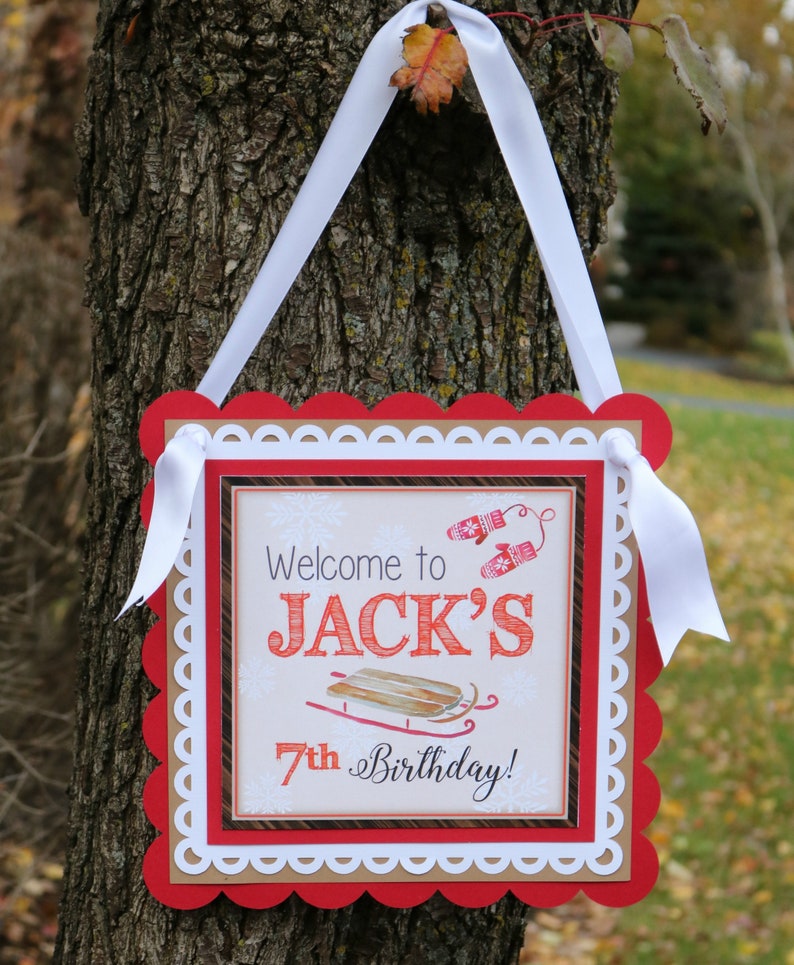 May include: A square sign with a white ribbon hanger featuring a red scalloped border and a white decorative edge. The center displays the text Welcome to JACK'S 7th Birthday! with illustrations of red mittens and a wooden sled on a light background.