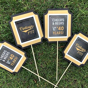 May include: Four black and gold square signs with white text. The signs say "Cheers & Beers to 40 Years!" and "Vintage 1977".
