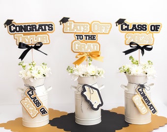 May include: Three graduation centerpiece decorations with gold glitter and black text. The centerpieces feature a milk jug vase with white flowers, a black and gold round placemat, and a graduation themed topper. The toppers read "Congrats Taylor", "Hats Off to the Grad", and "Class of 2024".