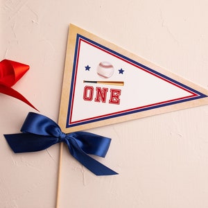 May include: A white pennant with a red, white, and blue border. The pennant has a baseball and bat graphic with the word "ONE" in red letters. The pennant is attached to a wooden stick with a blue ribbon bow.