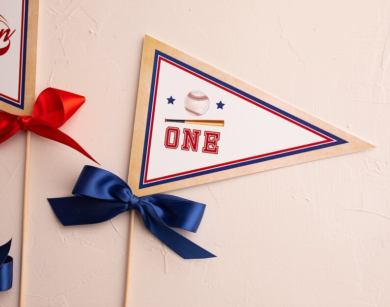 May include: A white pennant with a red, white, and blue border. The pennant has a baseball and bat graphic with the word "ONE" in red letters. The pennant is attached to a wooden stick with a blue ribbon bow.