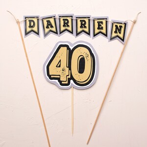 May include: A cake topper set featuring a banner that reads "DARREN" and a large number "40" in yellow with black outlines. The banner and number are attached to wooden sticks.