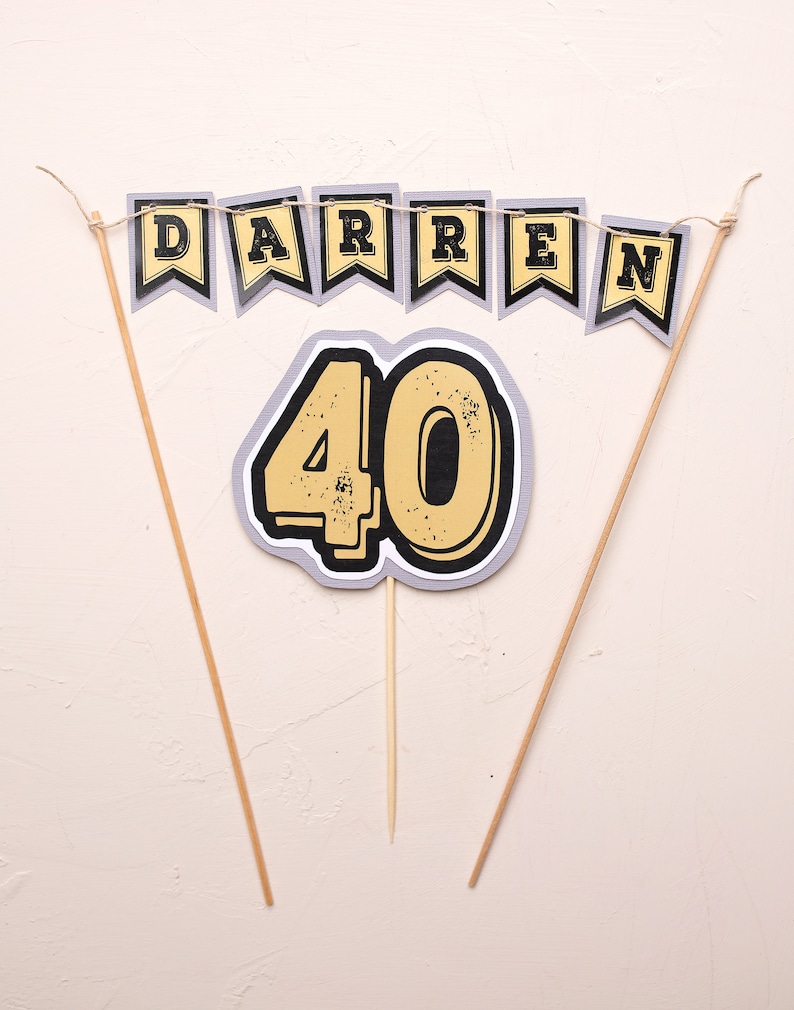 May include: A cake topper set featuring a banner that reads "DARREN" and a large number "40" in yellow with black outlines. The banner and number are attached to wooden sticks.