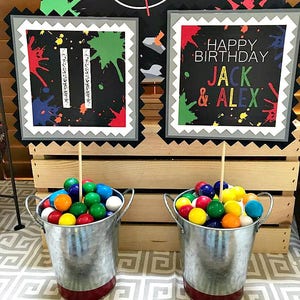 May include: Two small metal buckets filled with colorful gumballs. The buckets are decorated with a red stripe around the bottom.  Two signs with black backgrounds and colorful paint splatters are in the background. One sign says "Happy Birthday Jack & Alex". The other sign has the number "11" on it.