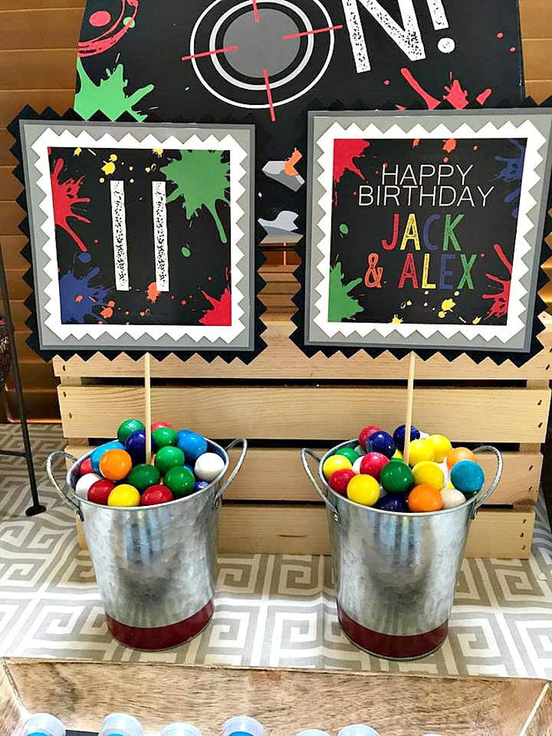 May include: Two small metal buckets filled with colorful gumballs. The buckets are decorated with a red stripe around the bottom.  Two signs with black backgrounds and colorful paint splatters are in the background. One sign says "Happy Birthday Jack & Alex". The other sign has the number "11" on it.