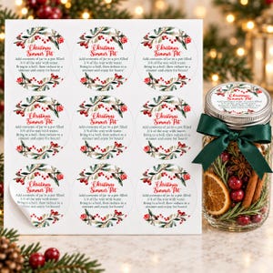 May include: A sheet of nine circular stickers and one glass jar filled with dried orange slices, cranberries, cinnamon sticks, and rosemary. Each sticker features a red and green holly illustration and the text Christmas Simmer Pot.