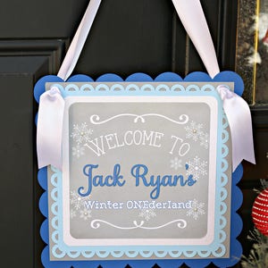 May include: A gray and blue welcome sign with white scalloped edges. The sign says "Welcome to Jack Ryan's Winter Wonderland".