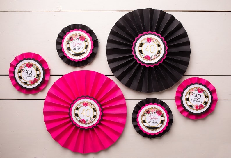 以下が含まれることがあります： 金色のアクセントと花柄のデザインが施された、黒とピンクの紙製扇風機6個。扇風機には、「Cheers to 40 Years」と「40 & Fabulous」の文字が印刷されています。