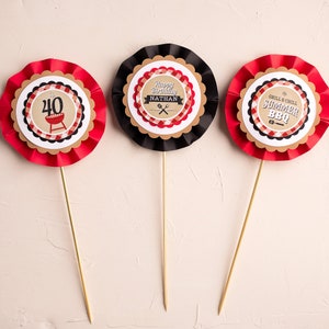 May include: Three red, white, and black paper cupcake toppers with a barbecue theme. The toppers feature a grill, a checkered pattern, and the text "Happy Birthday Nathan", "Grill & Chill Summer BBQ", and "40".