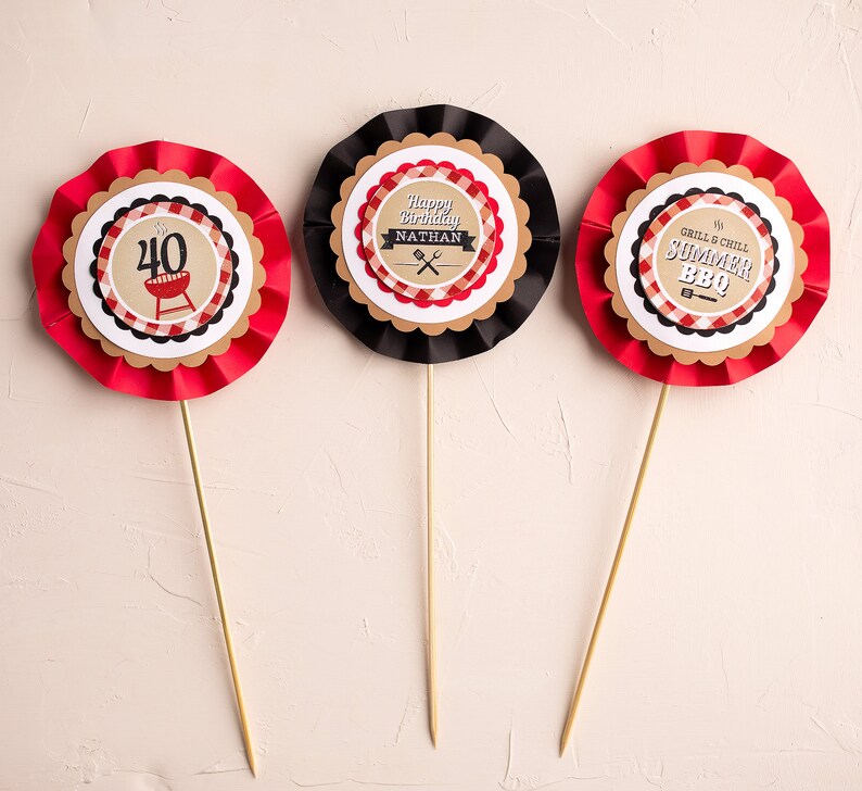 May include: Three red, white, and black paper cupcake toppers with a barbecue theme. The toppers feature a grill, a checkered pattern, and the text "Happy Birthday Nathan", "Grill & Chill Summer BBQ", and "40".