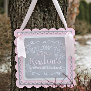 May include: A decorative sign with a scalloped pink border and a gray interior. The sign reads "Welcome to Keaton's Winter ONEDerland" in white script and snowflake accents. White ribbons are attached for hanging.