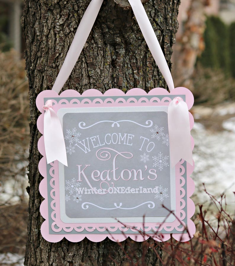May include: A decorative sign with a scalloped pink border and a gray interior. The sign reads "Welcome to Keaton's Winter ONEDerland" in white script and snowflake accents. White ribbons are attached for hanging.