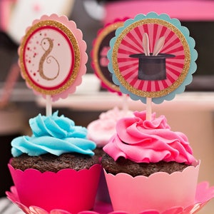 May include: Two cupcakes with pink and blue frosting on a pink cake stand. The cupcakes are decorated with a pink and gold cupcake topper with a magic hat and a pink and gold cupcake topper with the number 8.