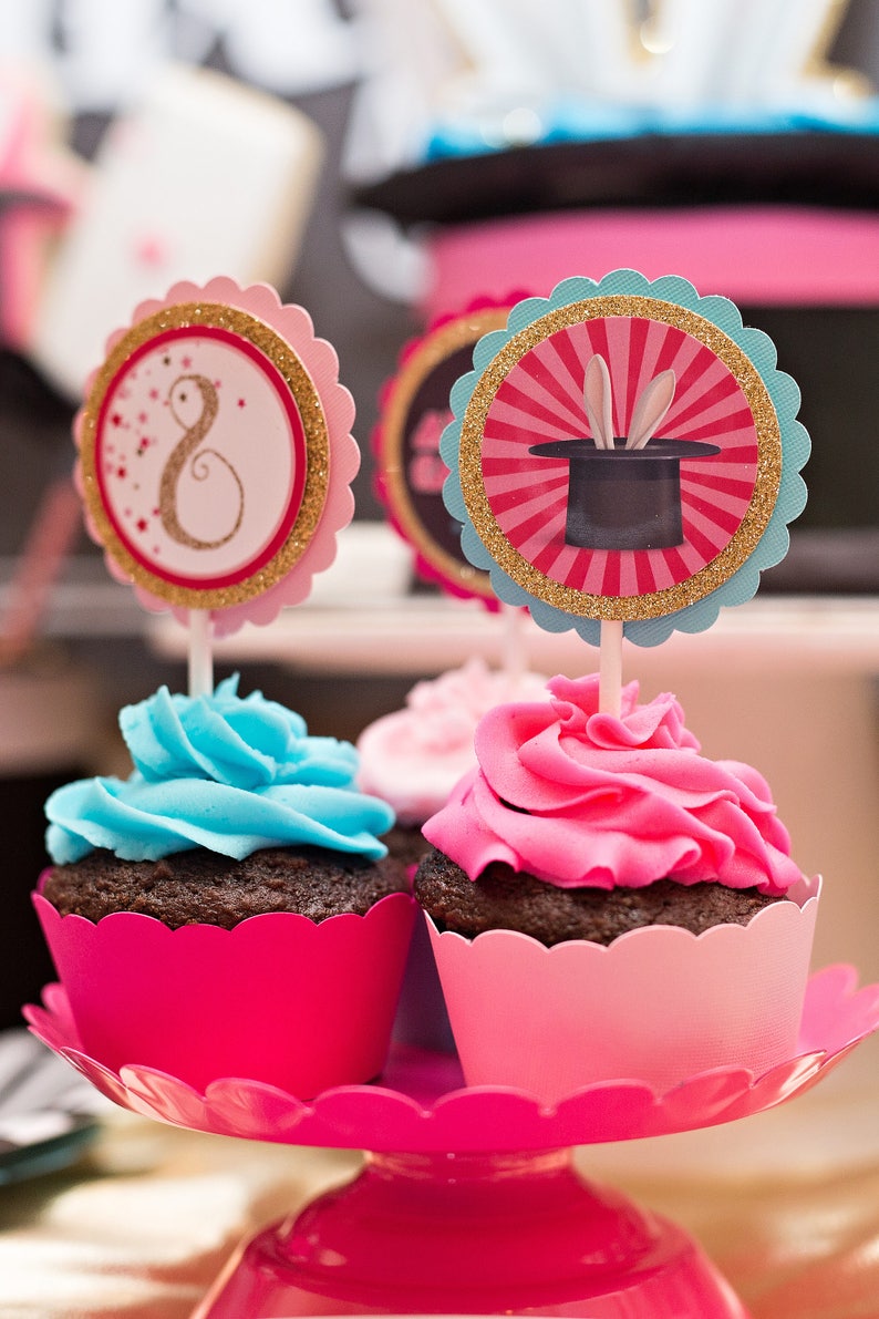 May include: Two cupcakes with pink and blue frosting on a pink cake stand. The cupcakes are decorated with a pink and gold cupcake topper with a magic hat and a pink and gold cupcake topper with the number 8.