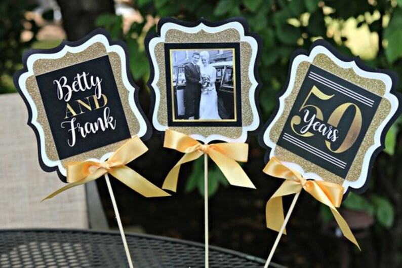 Puede incluir: Tres centros de mesa negros y dorados con lazos de cinta dorada. Los centros de mesa presentan una foto de una pareja, el texto "Betty y Frank" y el texto "50 a&ntilde;os".