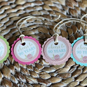 May include: Four colorful tags with scalloped edges and twine strings. Each tag has a white center with the text "cheers to 90 YEARS" and a floral design. The tags are in green, pink, light pink, and blue.
