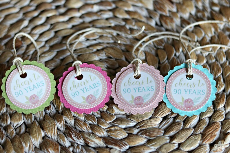 May include: Four colorful tags with scalloped edges and twine strings. Each tag has a white center with the text "cheers to 90 YEARS" and a floral design. The tags are in green, pink, light pink, and blue.