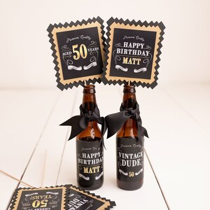 May include: Two brown glass bottles with black and gold labels that say "Happy Birthday Matt" and "Vintage Dude 50". The bottles are decorated with black ribbon and are topped with black and gold square signs that say "Aged 50 Years" and "Happy Birthday Matt".