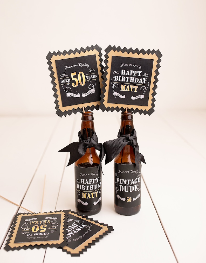 May include: Two brown glass bottles with black and gold labels that say "Happy Birthday Matt" and "Vintage Dude 50". The bottles are decorated with black ribbon and are topped with black and gold square signs that say "Aged 50 Years" and "Happy Birthday Matt".