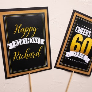 May include: Two black and gold birthday signs with white text. The first sign says "Happy Birthday Richard". The second sign says "Cheers to 60 Years".