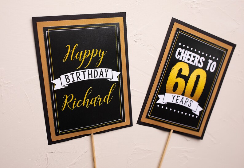 May include: Two black and gold birthday signs with white text. The first sign says "Happy Birthday Richard". The second sign says "Cheers to 60 Years".