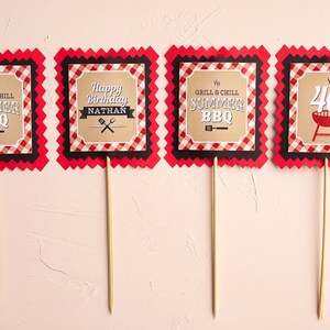 Puede incluir: Cuatro adornos para fiestas con bordes a cuadros rojos y negros. Los adornos tienen texto que dice "GRILL & CHILL SUMMER BBQ", "Happy Birthday NATHAN" y "40" con un gr&aacute;fico de barbacoa. Los adornos est&aacute;n sobre palitos de madera.