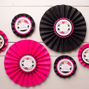 May include: A collection of decorative paper fans in black and hot pink, with a white wooden background. Each fan features a central circular design with the number "40" and floral accents, with text that reads "Cheers to 40 Years" and "40 & Fabulous".
