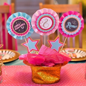 May include: A pink and blue table centerpiece with three paper rosettes, each with a different name, "Anna Gabriela", "Rose", and a number "8". The rosettes are attached to sticks and placed in a gold glitter top hat with pink tissue paper. The centerpiece is surrounded by gold glitter and pink and blue stars.