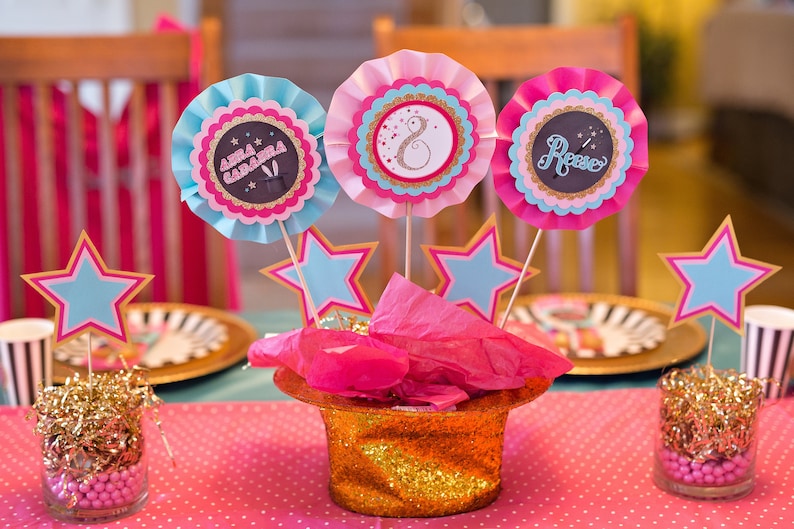 May include: A pink and blue table centerpiece with three paper rosettes, each with a different name, "Anna Gabriela", "Rose", and a number "8". The rosettes are attached to sticks and placed in a gold glitter top hat with pink tissue paper. The centerpiece is surrounded by gold glitter and pink and blue stars.