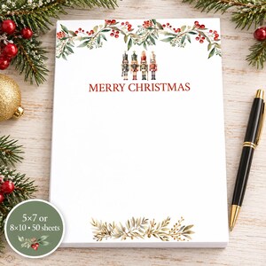 May include: A white rectangular notepad featuring a watercolor illustration of four nutcrackers and red berries with green leaves at the top. The text MERRY CHRISTMAS is printed in red below the nutcrackers. A green circular label states 5x7 or 8x10 50 sheets.