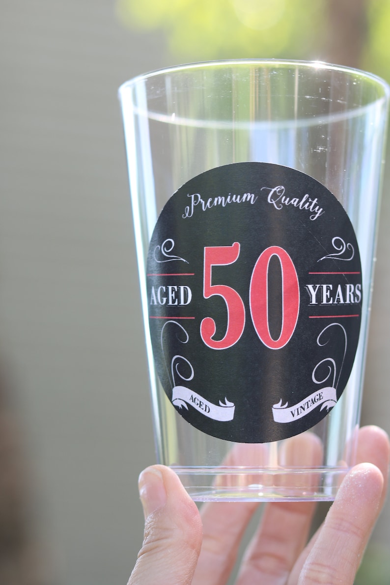 May include: A clear plastic cup with a black and white sticker that says "Premium Quality Aged 50 Years Vintage". The number 50 is in red.